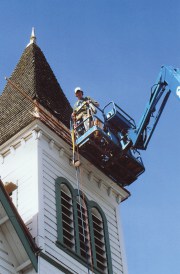 man standing in blue crane with hard hat looks down from the side of a church steeple. It is a tower white with green trim and grey roofing.