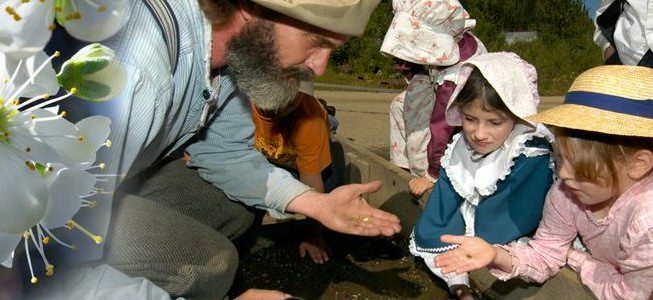 Bearded man and two children examining a coin. All are wearing historic attire.