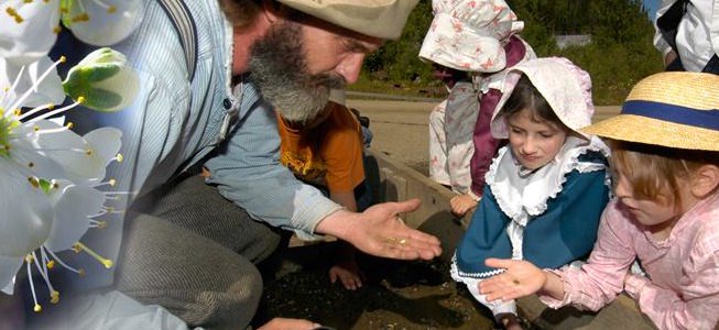 Bearded man and two children examining a coin. All are wearing historic attire.