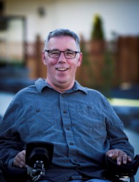 White middle aged man with glasses wearing a blue button up shirt smiles for the camera. He is seated in a wheelchair.