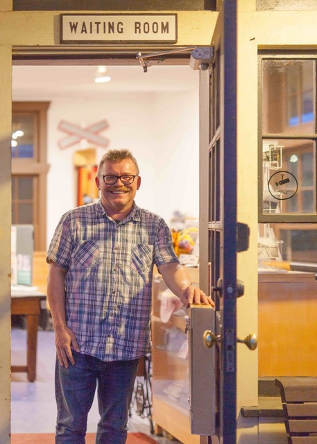 white man stands inside doorway. He's wearing glasses and a collared button-up tee-shirt. He is smiling.