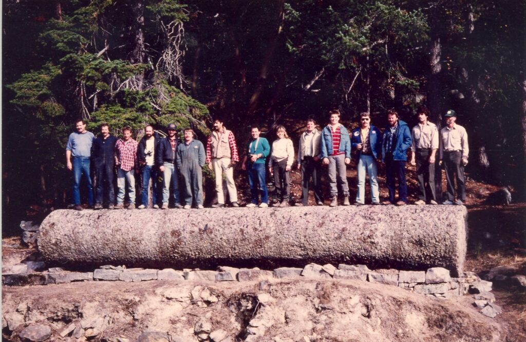 15 adults pose on top of a large wooden column from the shipwrecked Zephyr laying on its side.