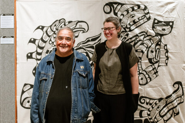 Middle aged Indigenous man wearing a black shirt and jean jacket strands to the left of a middle aged white woman with grey hair wearing classes and a green blouse. Behind them is a large piece of hanging art.