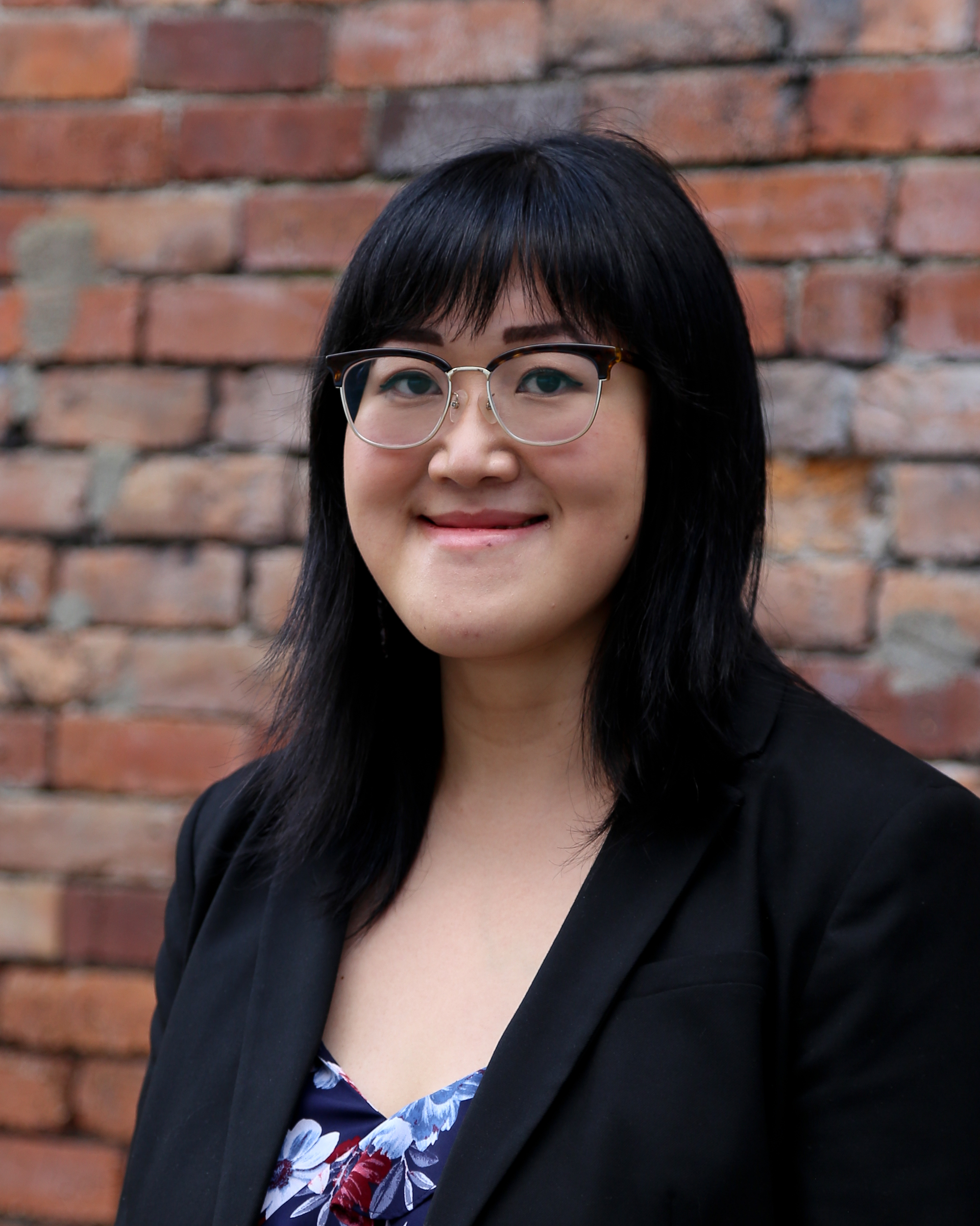 Chinese Canadian woman with mid-length black hair and bangs wearing a glasses, a black blazer and a patterned blouse