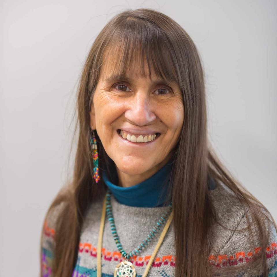 middle aged Indigenous woman with long light brown hair and bangs swearing a teal turtleneck and sweater with colourful stripes