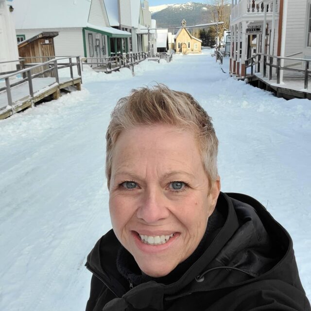 white woman with short blond hair smiling in a black winter coat ground behind her is snowy