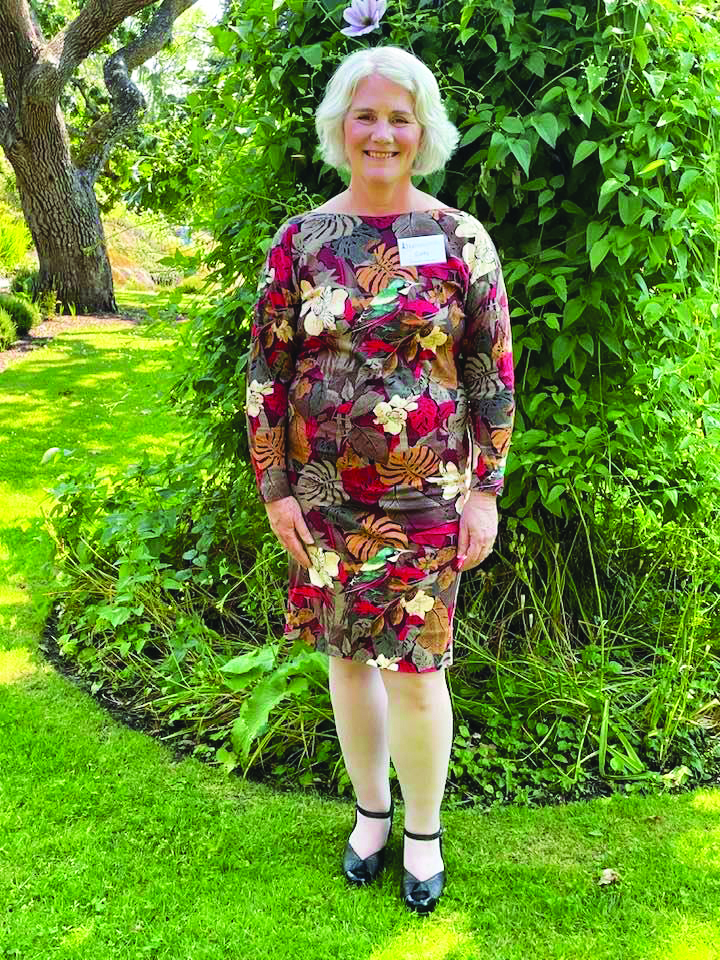 Senior white woman with short white hair poses on grass in front of a tree wearing a floral patterned dress and black shoes