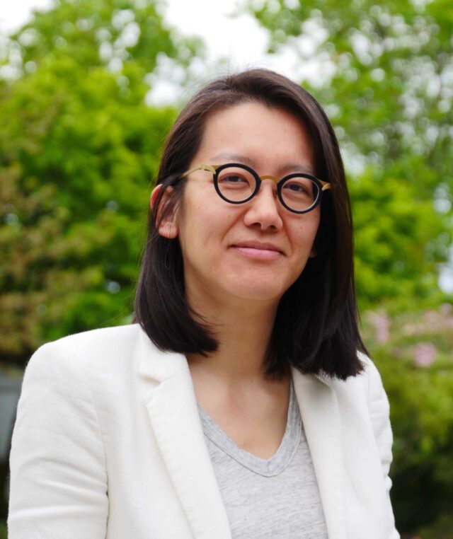 A Chinese Canadian woman with midlength black hair wearing a grey shirt, white blazer and black rimmed round glasses.