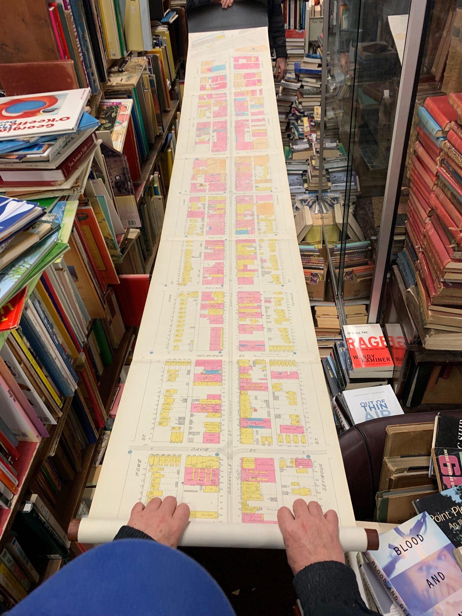 A scroll document rolled out shows a city map of blocks with buildings coloured in pink or yellow. Map is surrounded by stacked books everywhere.