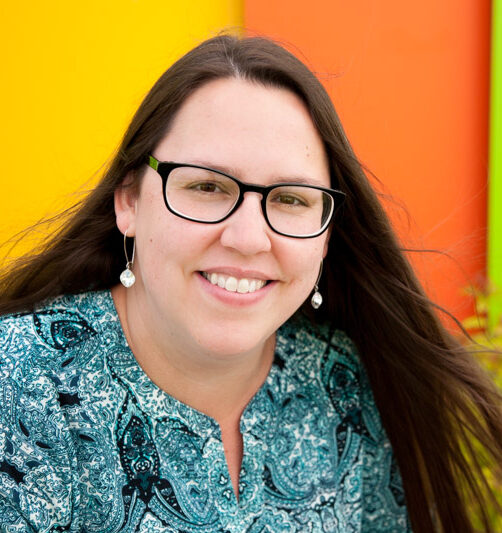 indigenous woman with long brown hair wearing black framed glasses and a patterned teal shirt smiling in front of a brightly coloured wall