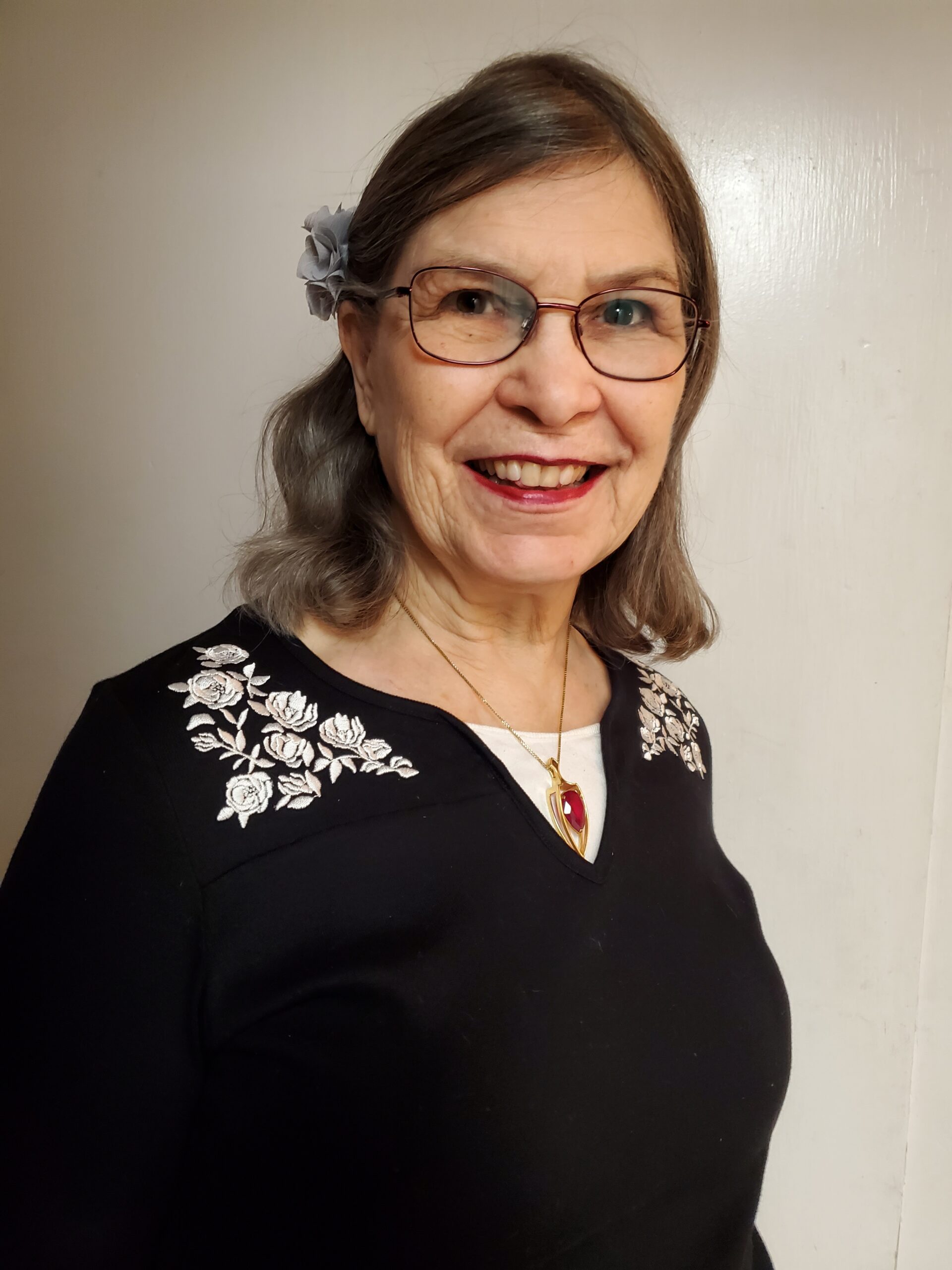 headshot of a senior white woman with medium length greying hair wearing glasses and a flower in her hair. She is smiling.