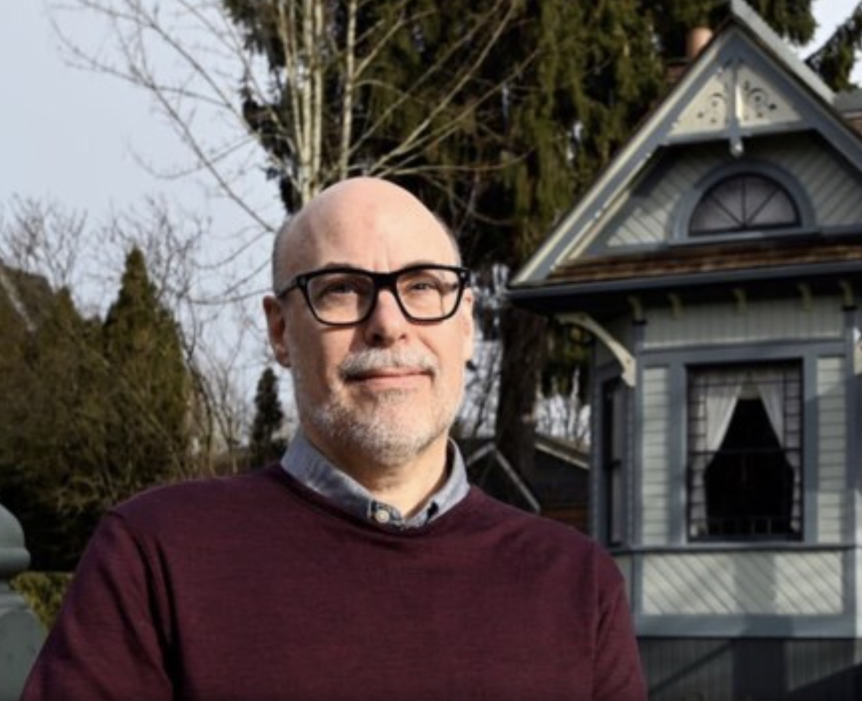 middle aged bald man with mustache and beard wearing black acetate glasses and a maroon sweater with collared shirt. Posing in front of an historic house and trees.