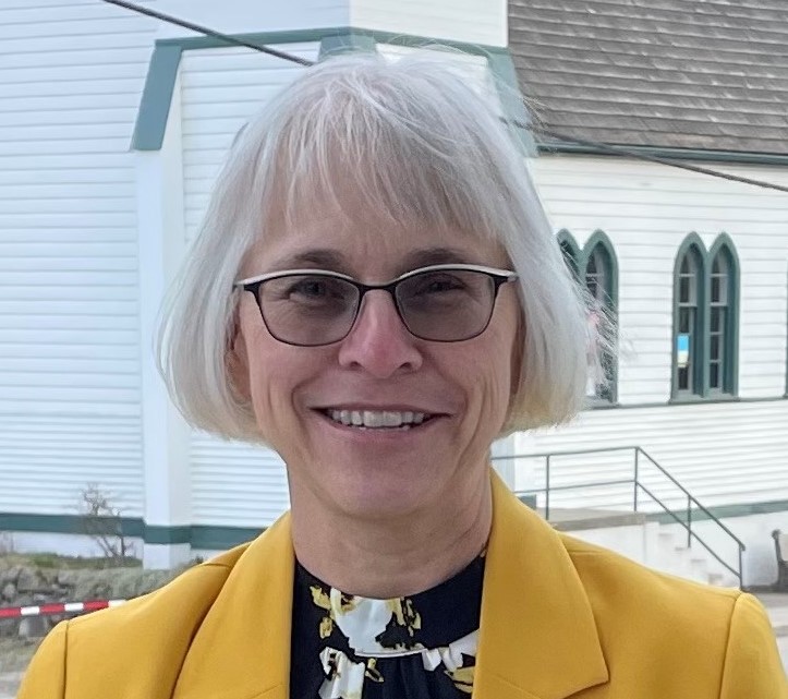 White haired with bangs middle aged white woman wearing glasses and a yellow blazer