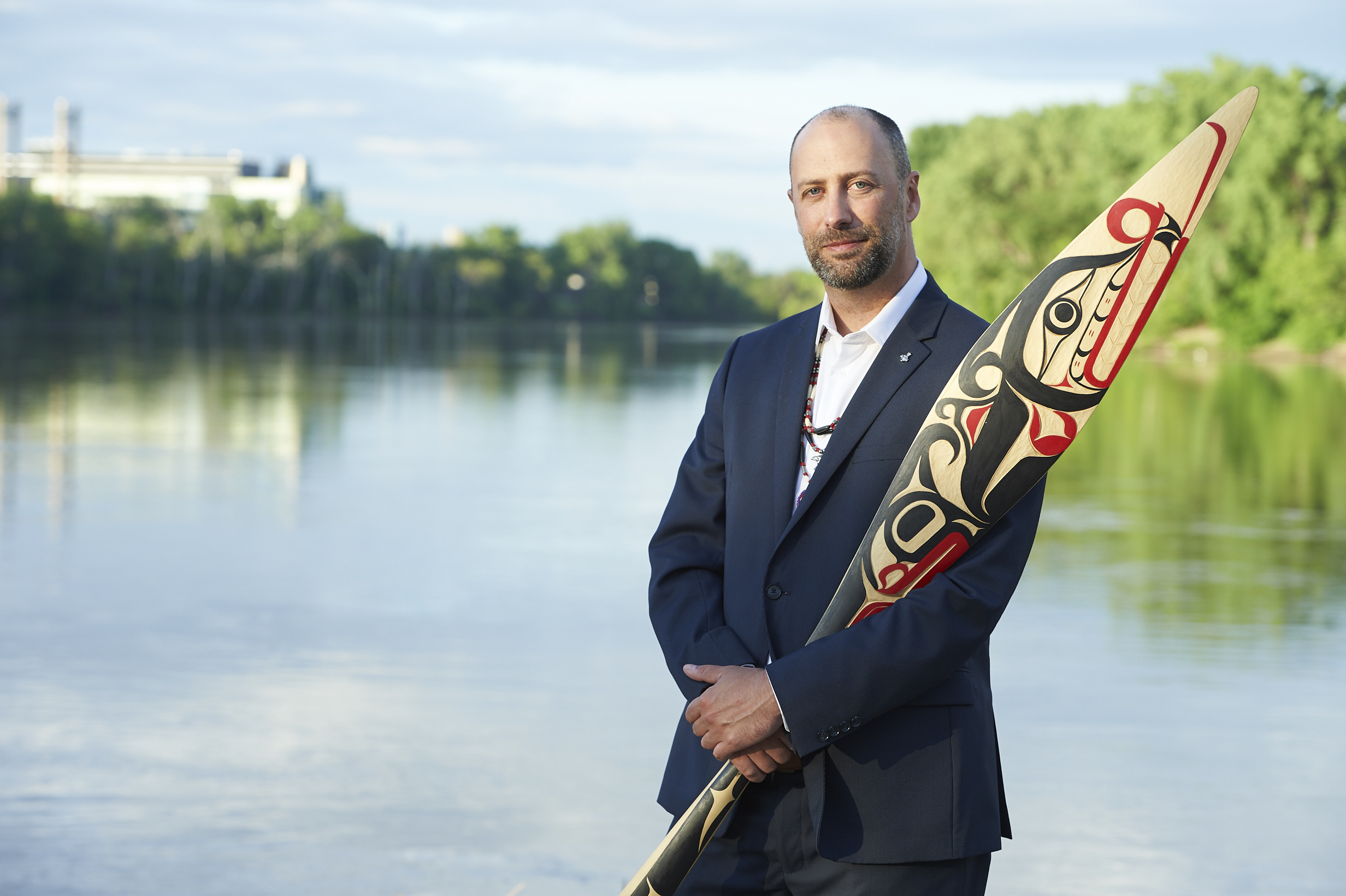 Indigenous man with a beard wearing a suit poses with a wooden paddle painted in black and red.
