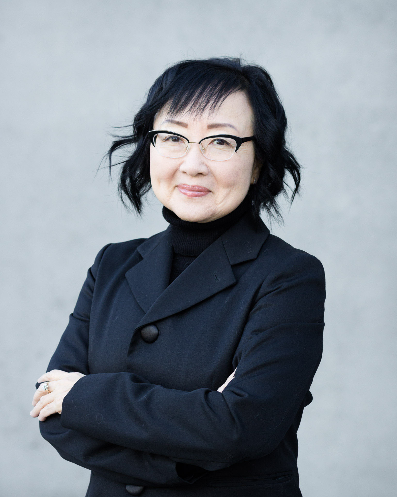 A middle aged Japanese Canadian woman with short wavy black hair wearing black rimmed glasses and a black blazer and black turtleneck.