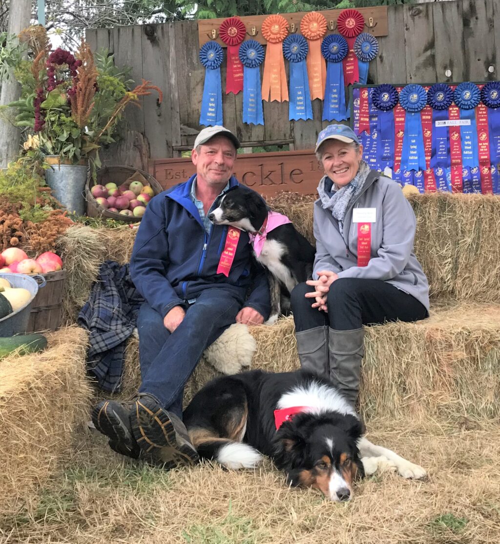 Marjorie and Mike sit on hay stacks wearing coats and baseball caps. They are both wearing ribbon prizes and dozens more ribbons are hanging on the barn wall behind them.