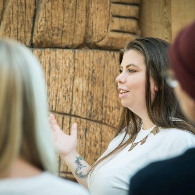 Young indigenous woman with long brown hair wearing a white sweater and necklace speaking in front of a stone wall
