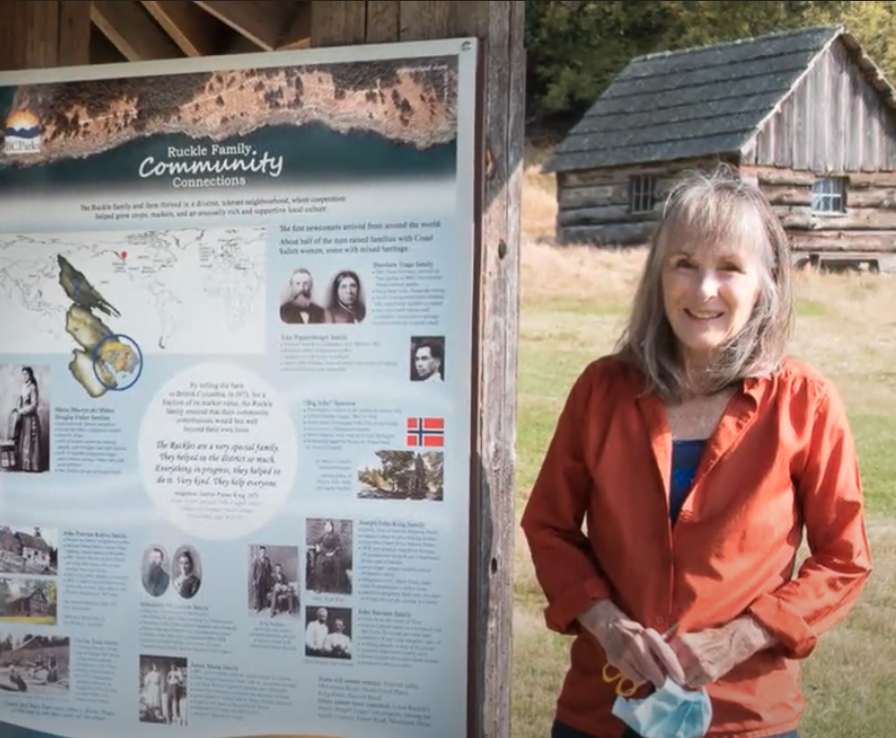 grey haired woman in an orange rain jacket stands in front of an interpretive sign for Ruckle Farm