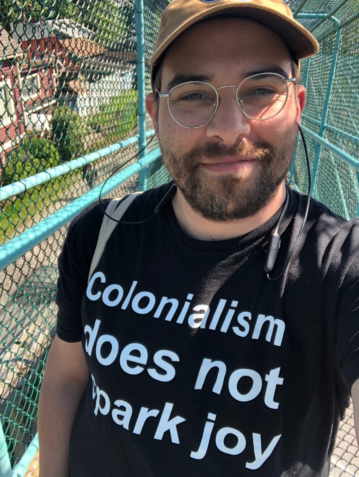 non-binary person with a brown beard wearing a baseball hat, glasses and a tee-shirt that reads 'colonialism does not spark joy'
