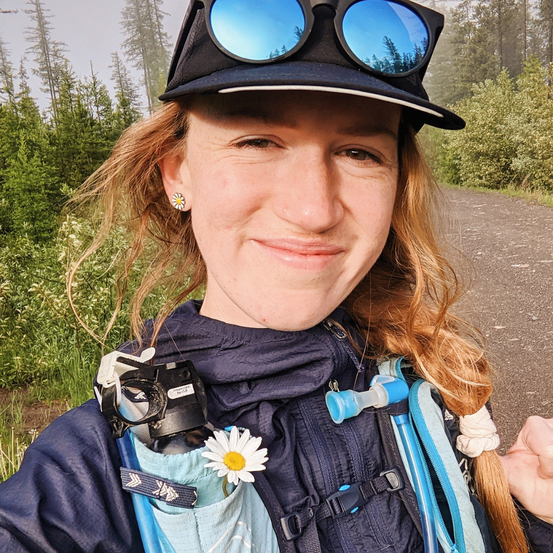 Jennyce has fair skin and long light brown hair in a braid under a baseball hat. She is on a hike and the photo is cropped to just her face and shoulders.