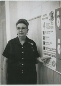 black and white photo of Ruby Nobbs when she was middle aged. She wears a dark bowling shirt and glasses. She is pointing at Youth Bowlers Championships Poster