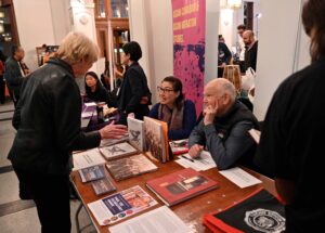 two people sit at a table covered with documents at books they speak with a woman standing on the other side of the table 