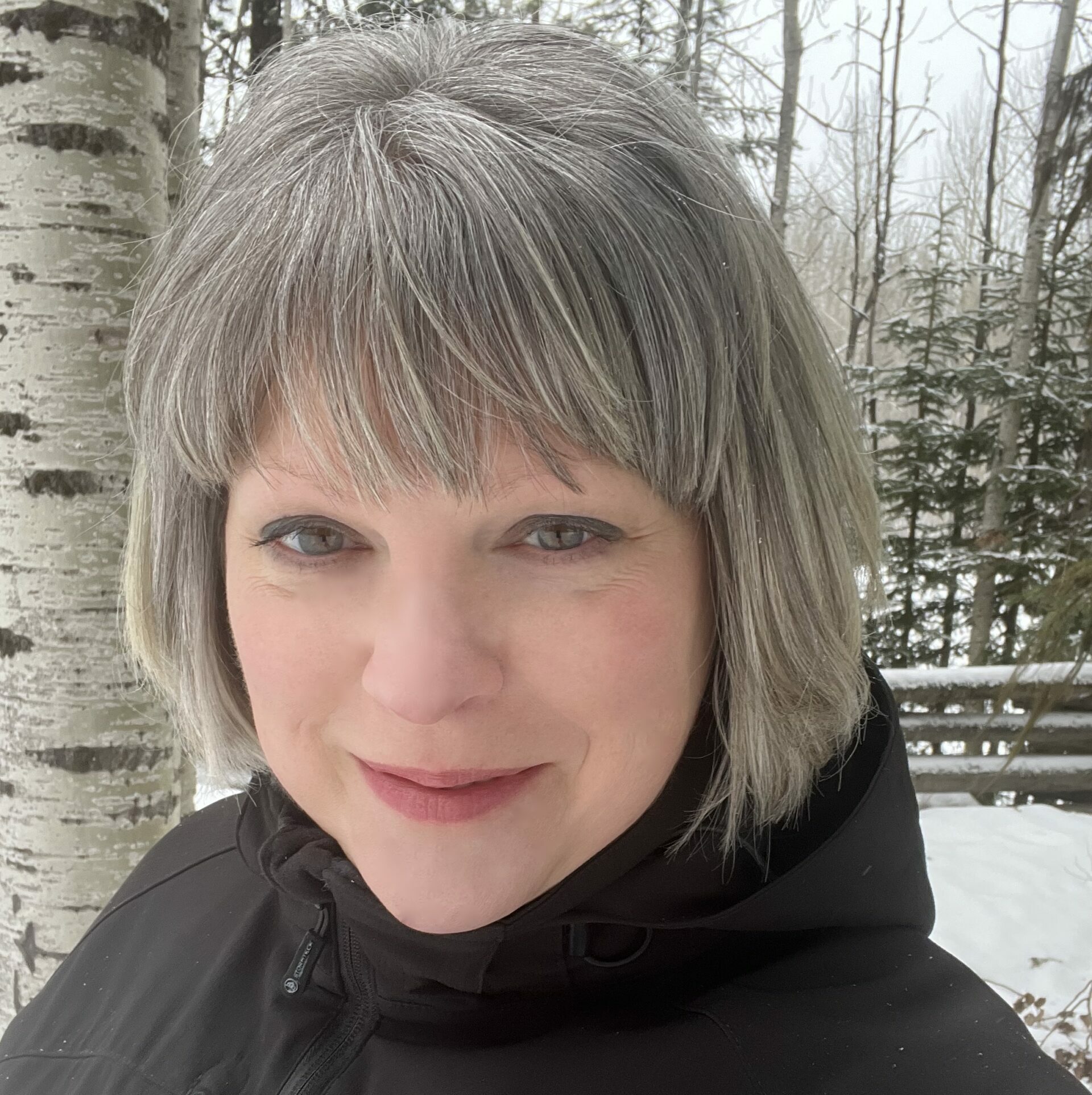 headshot of smiling grey haired woman wearing a black jacket that has "Barkerville" embroidered on the chest standing outside in the woods.