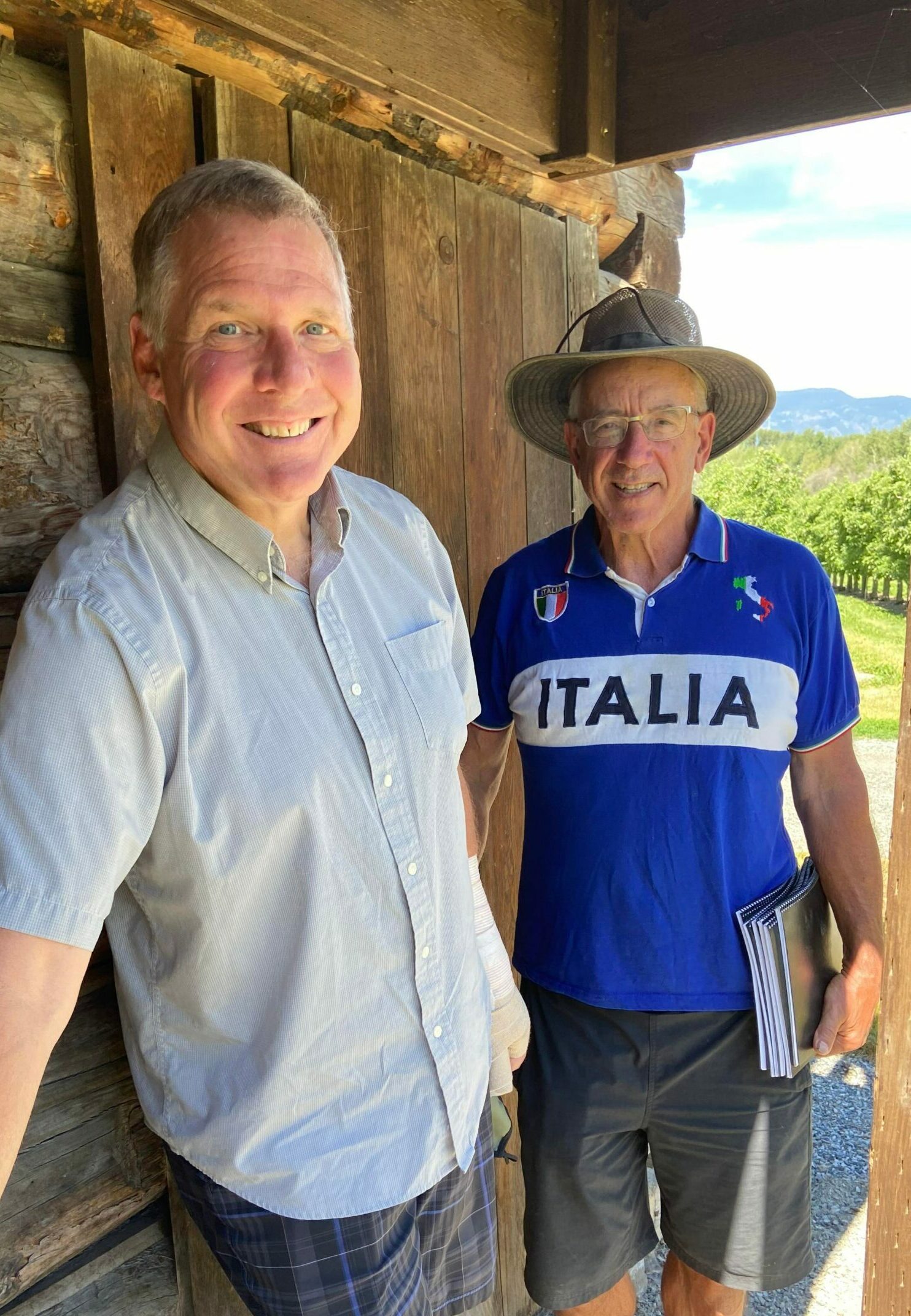 two senior men with white hair standing outside a log cabin.