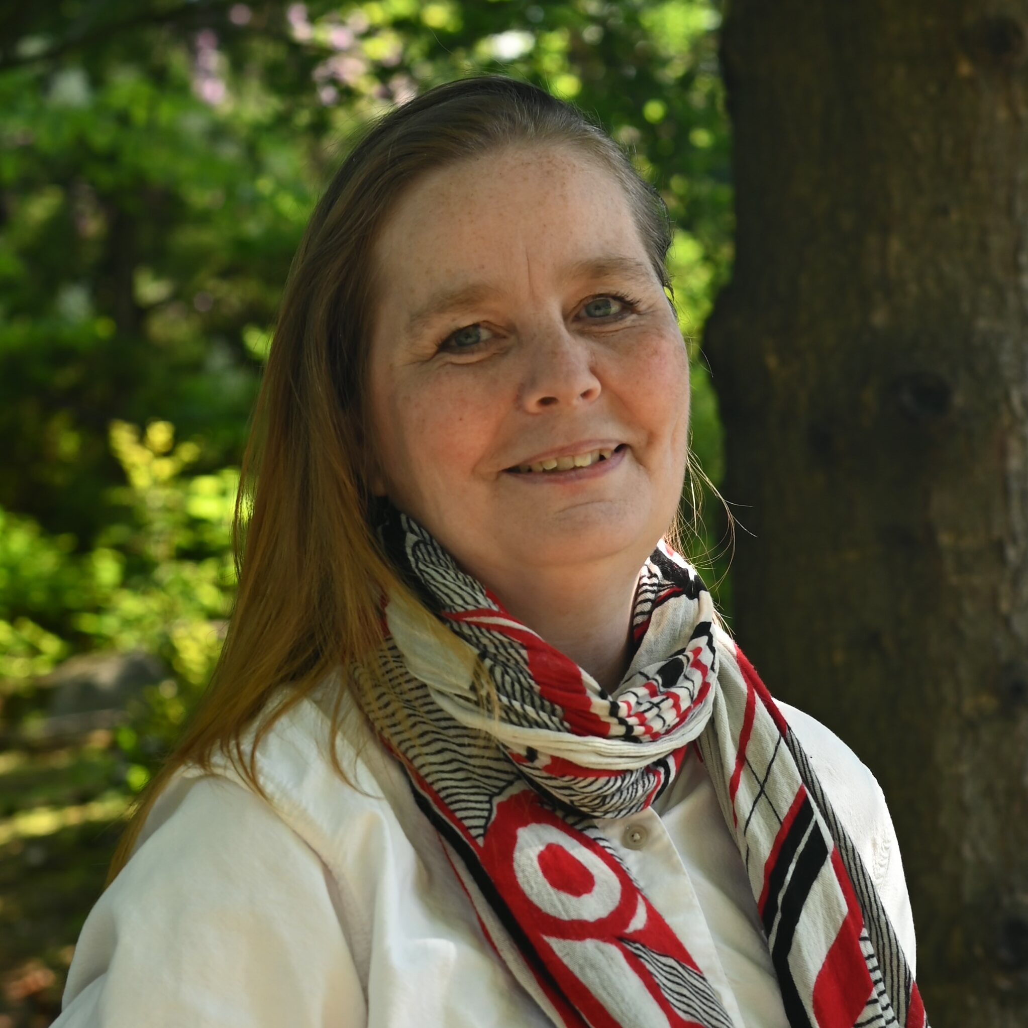 white woman with long hair wearing a white sweater and patterned scarf posing in front of a tree in a park