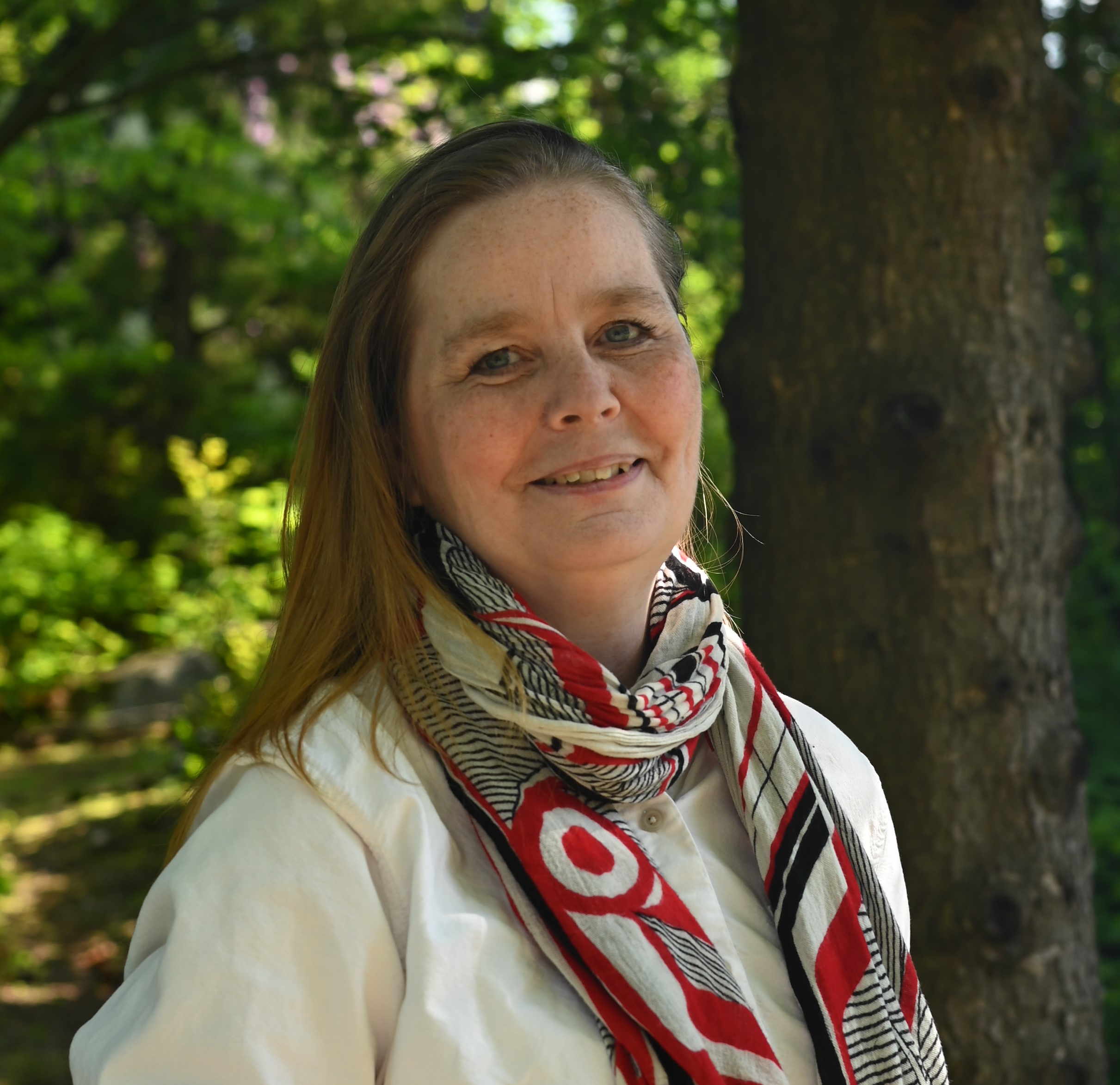 white woman with long hair wearing a white sweater and patterned scarf posing in front of a tree in a park