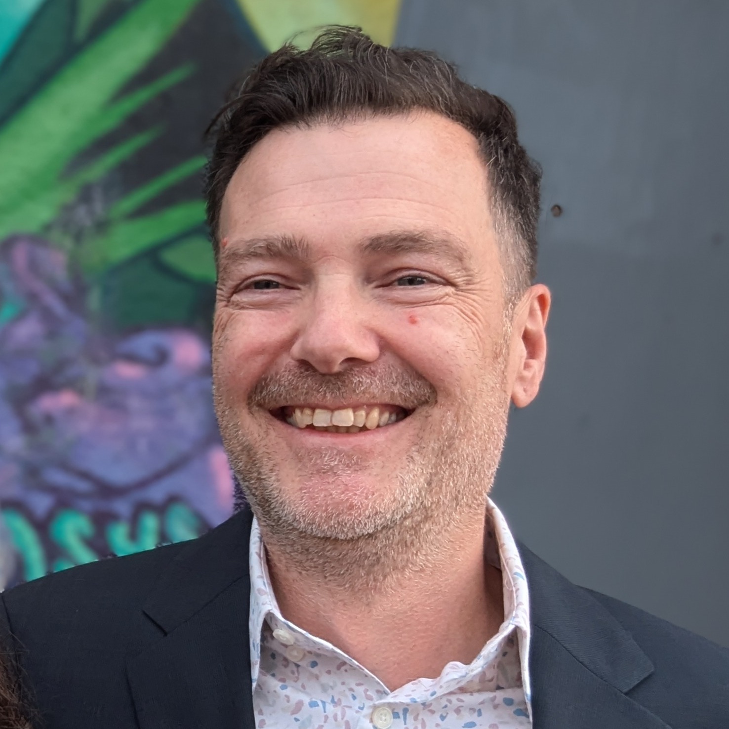 Daytime, outdoors, a very handsome man looking slightly up and left, smiling wide. He is wearing a blue suit over a pastel terra-cotta patterned shirt. Behind him is a concrete wall with part of a graffiti mural visible.